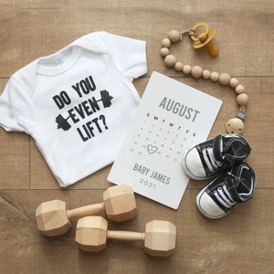May include: A white babygrow with the text "Do you even lift?" and a black dumbbell graphic. A calendar with the month of August and the name "Baby James" is on a wooden surface. There are two wooden dumbbells, a dummy, and a pair of baby shoes.