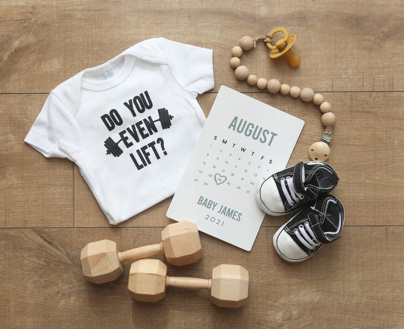 May include: A white babygrow with the text "Do you even lift?" and a black dumbbell graphic. A calendar with the month of August and the name "Baby James" is on a wooden surface. There are two wooden dumbbells, a dummy, and a pair of baby shoes.