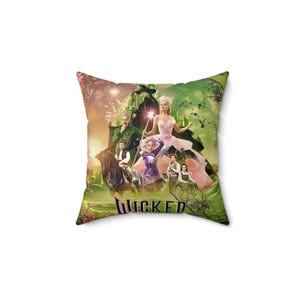 Peut inclure: Un coussin vert et rose avec un motif représentant la distribution de la comédie musicale Wicked. Le coussin présente les personnages d'Elphaba, Glinda, Fiyero et le Magicien. Le texte "WICKED" est imprimé sur le coussin.