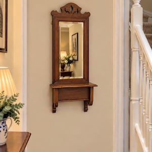 May include: A rectangular wall-mounted mirror with a dark brown wooden frame and a decorative carved crest at the top. The mirror features an integrated small wooden shelf at the base, supported by two carved brackets, providing a functional ledge.