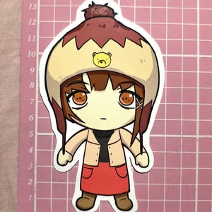 May include: A sticker featuring a cartoon character with brown hair and large orange eyes. The character wears a tan jacket, red skirt, and a brown hat with a bear design. The sticker is on a pink grid background.