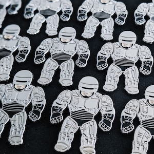 Może przedstawiać: Kolekcja srebrnych pinów z robotami. Każdy pin przedstawia robota w srebrnym hełmie i z detalicznym korpusem. Roboty stoją w różnych pozach.