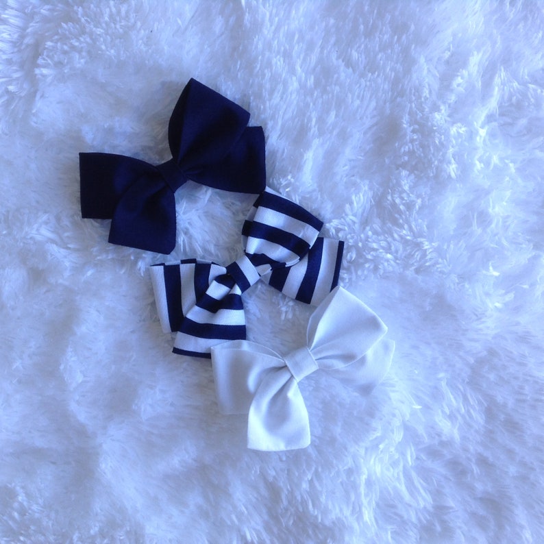 May include: Three hair bows in navy, white, and navy and white striped patterns. The bows are made of fabric and are arranged on a white, fluffy surface. The bows are a classic accessory.