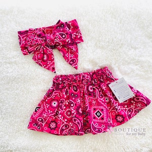 May include: Pink and black bandana print baby skirt and matching headband. The skirt has a gathered elastic waistband and the headband is tied in a bow.
