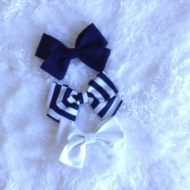 May include: Three fabric hair bows in a top-down view. One is navy blue, one is white with navy blue stripes, and the last is solid white. The bows are arranged on a fluffy white surface.