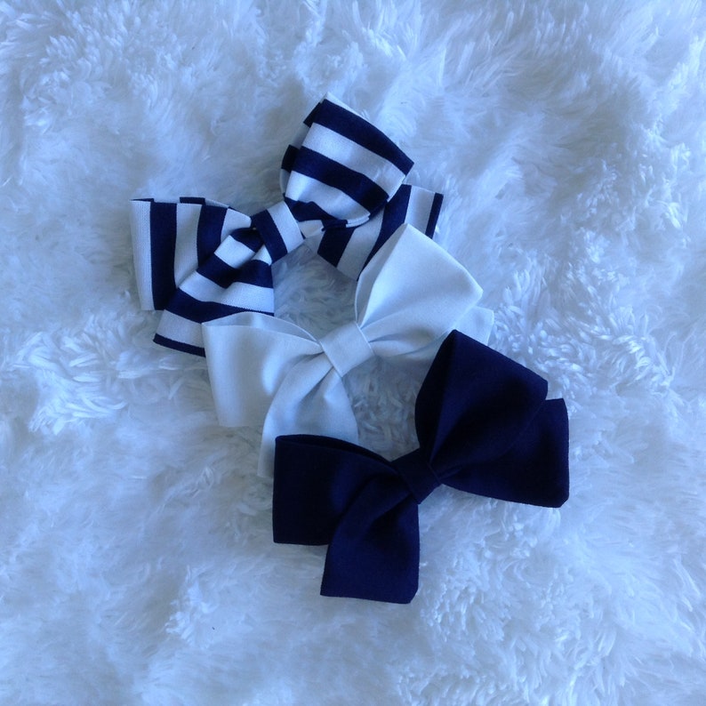 May include: Three hair bows in a close-up shot. One bow is navy blue and solid, another is white, and the third is white with navy blue stripes. The bows are arranged on a fluffy white surface.