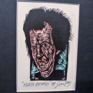 May include: A framed woodcut print of a man's portrait. The artwork features a black and pink color scheme, with the man's face and hair rendered in intricate detail. The text "Keith Richards" AIP Sean Eary is visible.