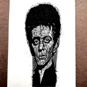 May include: Black and white linocut portrait of a man with dark, curly hair and a stern expression. The artwork features detailed cross-hatching and the text "LOU REED A/P SEAN EABY" at the bottom. The portrait is on a white background.