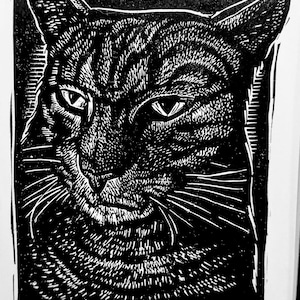 May include: Black and white linocut print of a tiger cat's face. The cat has detailed fur patterns, intense eyes, and long whiskers. The print is signed "Sean Eaby" and titled "Tiger Cat" with "A/P" below the title.