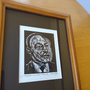 May include: Framed black and white portrait of Martin Luther King Jr. The artwork is a linocut print with detailed lines and shading, set against a black background. The frame is made of wood.