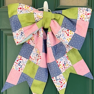 May include: A large, decorative bow made of patchwork fabric, featuring squares of pink, green, and blue with floral and polka dot patterns. The bow is hanging on a green door, adding a touch of handmade charm.