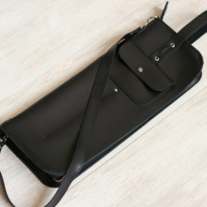 K&ouml;nnte beinhalten: Eine schwarze Leder-Umh&auml;ngetasche mit einer gro&szlig;en Klappe und einer kleineren Rei&szlig;verschlusstasche. Die Tasche hat einen langen, verstellbaren Riemen.