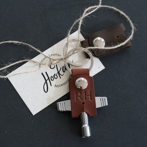 May include: Brown leather key chain with a silver key and the initials "RH" engraved on the leather. The keychain is attached to a silver key ring. The keychain is accompanied by a small brown leather tag with the text "Handmade Leather Hookah Accessories".