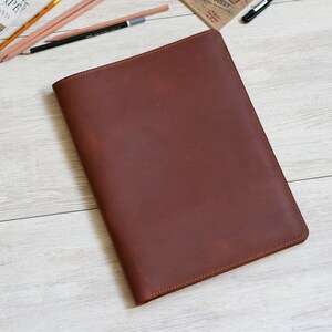 May include: A brown leather portfolio folder with a stitched edge. The folder is lying flat on a light wood surface.