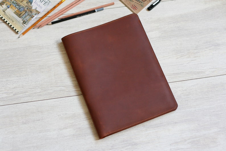 May include: A brown leather portfolio folder with a stitched edge. The folder is lying flat on a light wood surface.