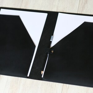 May include: A black leather folder with two pockets and a pencil holder. The folder is open and has two white sheets of paper inside.