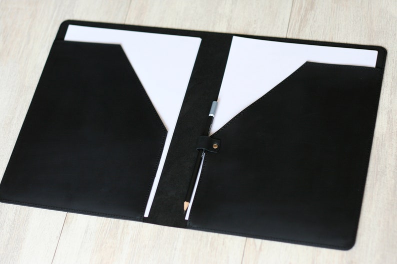 May include: A black leather folder with two pockets and a pencil holder. The folder is open and has two white sheets of paper inside.