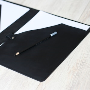 May include: A black leather folder with a white paper insert and a pencil inside. The folder has a flap that closes with a snap.