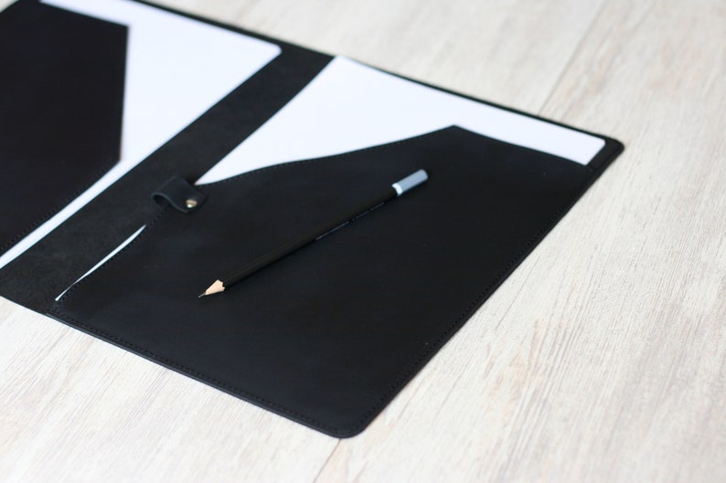 May include: A black leather folder with a white paper insert and a pencil inside. The folder has a flap that closes with a snap.