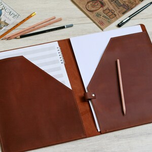 May include: A brown leather folder with two pockets and a flap closure. The folder is open and has a sheet of music paper and a blank sheet of paper inside. A pencil is sticking out of the right pocket.