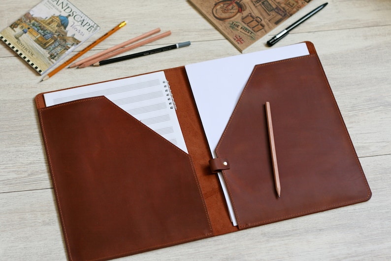 May include: A brown leather folder with two pockets and a flap closure. The folder is open and has a sheet of music paper and a blank sheet of paper inside. A pencil is sticking out of the right pocket.