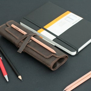 May include: A brown leather pencil case with a strap and a pencil sticking out. The case is rolled up and sitting on a black surface. There are also two pencils and a red pen on the surface.