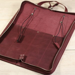 K&ouml;nnte beinhalten: Eine bordeauxrote Ledertasche mit Rei&szlig;verschluss und zwei Riemen mit Schnallen. Die Tasche ist offen und leer.
