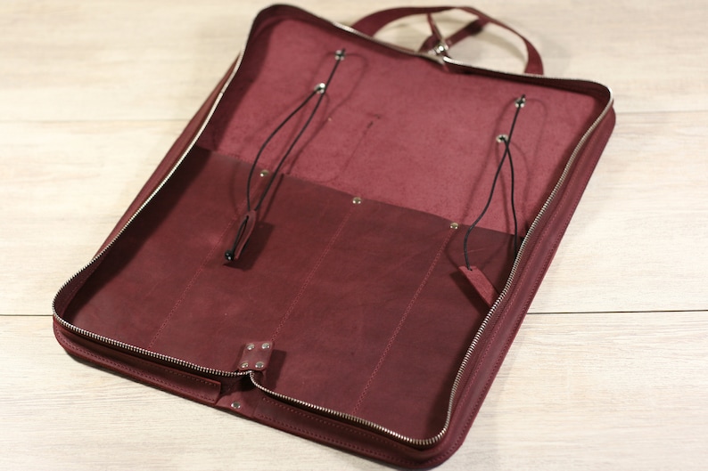 K&ouml;nnte beinhalten: Eine bordeauxrote Ledertasche mit Rei&szlig;verschluss und zwei Riemen mit Schnallen. Die Tasche ist offen und leer.