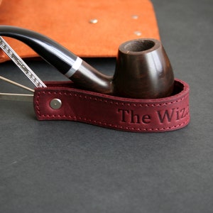 Puede incluir: Un porta pipas de cuero burdeos con el texto "The Wiz" grabado en él. Una pipa de madera descansa en el porta pipas.