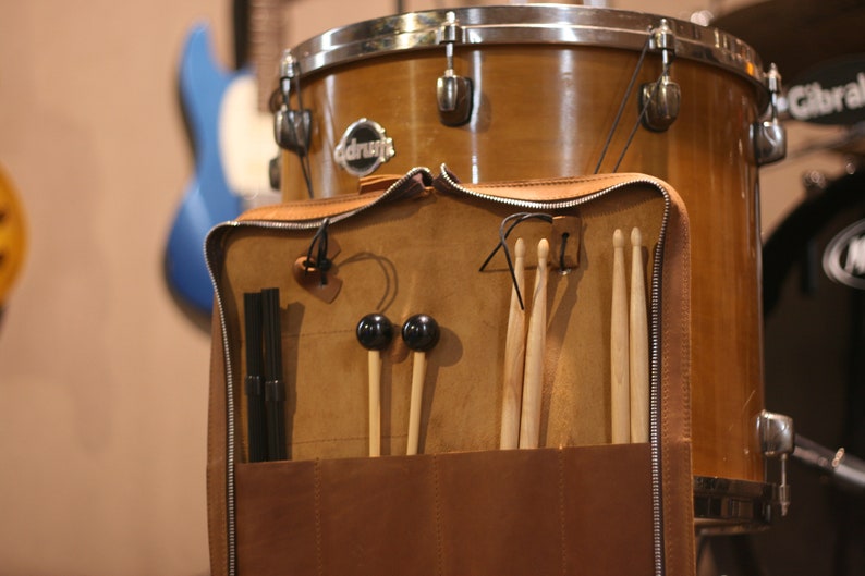 K&ouml;nnte beinhalten: Eine braune Ledertrommeltasche mit Rei&szlig;verschluss. Die Tasche hat mehrere F&auml;cher zum Verstauen von Drumsticks, Schl&auml;geln und anderem Zubeh&ouml;r. Die Tasche ist an einer braunen Trommel mit silbernem Rand befestigt, auf der das Wort "drumcraft" aufgedruckt ist.