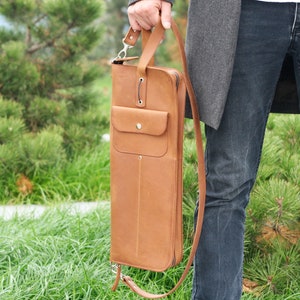 K&ouml;nnte beinhalten: Eine braune Ledertasche mit einem langen Riemen und einer kleinen Tasche auf der Vorderseite. Die Tasche wird von einer Person getragen, die Jeans und einen grauen Mantel tr&auml;gt.