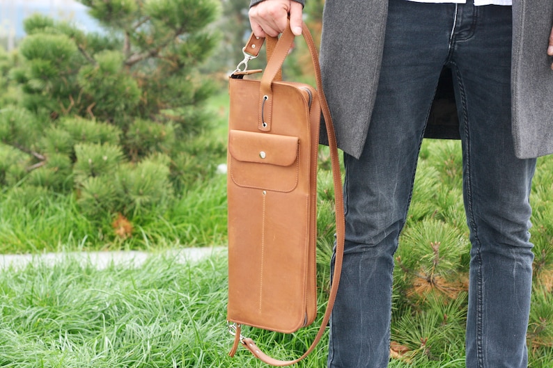 K&ouml;nnte beinhalten: Eine braune Ledertasche mit einem langen Riemen und einer kleinen Tasche auf der Vorderseite. Die Tasche wird von einer Person getragen, die Jeans und einen grauen Mantel tr&auml;gt.