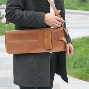 K&ouml;nnte beinhalten: Eine braune Ledertasche mit einem Klappverschluss und einer kleinen Tasche vorne. Die Tasche wird &uuml;ber der Schulter getragen und hat einen Riemen.