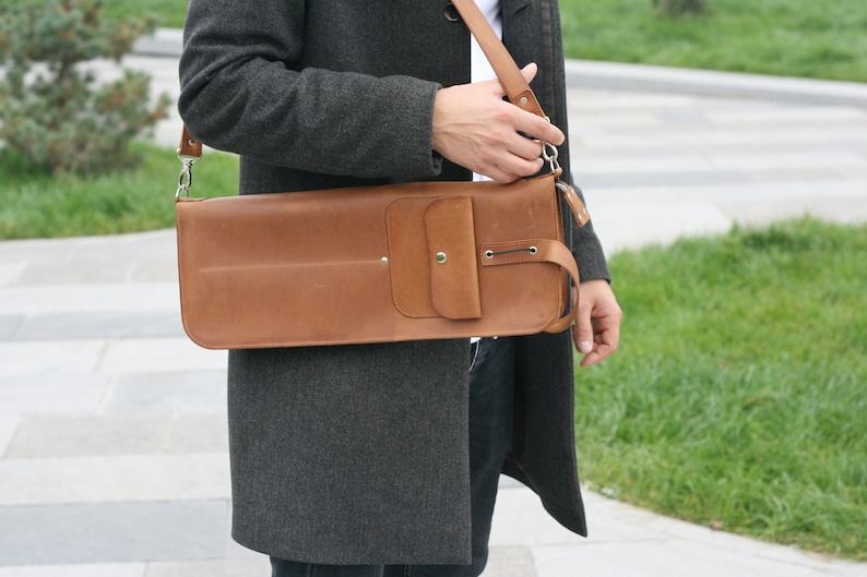 K&ouml;nnte beinhalten: Eine braune Ledertasche mit einem Klappverschluss und einer kleinen Tasche vorne. Die Tasche wird &uuml;ber der Schulter getragen und hat einen Riemen.