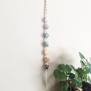 May include: A macrame wall hanging with seven chakra crystals in a rainbow colour scheme. The crystals are white, purple, blue, green, yellow, orange, and black. The macrame is made of natural white cord.