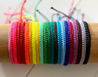 May include: A collection of colorful woven friendship bracelets in various shades of pink, red, orange, yellow, green, blue, purple, and gray. The bracelets are arranged in a rainbow pattern and are displayed on a wooden surface.
