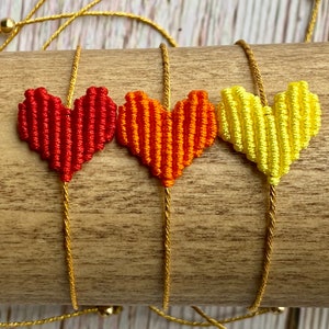 Puede incluir: Tres pulseras con dijes en forma de coraz&oacute;n hechos de hilo tejido en rojo, naranja y amarillo. Cada coraz&oacute;n est&aacute; unido a un cord&oacute;n dorado met&aacute;lico fino que se extiende verticalmente, con peque&ntilde;as cuentas doradas visibles en los extremos.