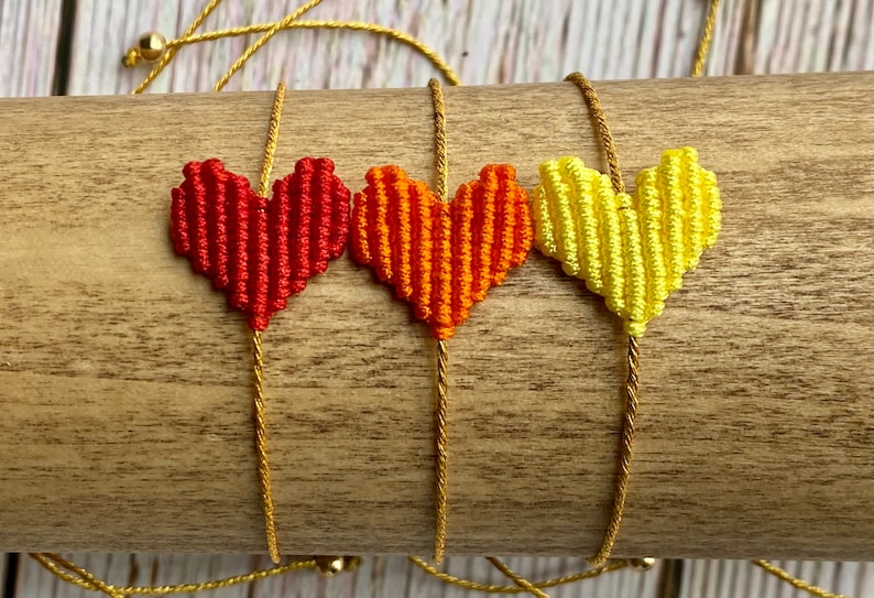 Puede incluir: Tres pulseras con dijes en forma de coraz&oacute;n hechos de hilo tejido en rojo, naranja y amarillo. Cada coraz&oacute;n est&aacute; unido a un cord&oacute;n dorado met&aacute;lico fino que se extiende verticalmente, con peque&ntilde;as cuentas doradas visibles en los extremos.