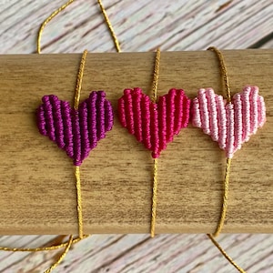 Puede incluir: Tres pulseras con dijes en forma de coraz&oacute;n hechos de hilo tejido en morado, magenta y rosa claro. Cada coraz&oacute;n est&aacute; centrado en un cord&oacute;n fino de color dorado met&aacute;lico. Las pulseras se muestran horizontalmente, destacando los colores vibrantes y la construcci&oacute;n texturizada y anudada de las piezas centrales en forma de coraz&oacute;n.