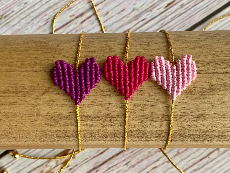 Puede incluir: Tres pulseras con dijes en forma de coraz&oacute;n hechos de hilo tejido en morado, magenta y rosa claro. Cada coraz&oacute;n est&aacute; centrado en un cord&oacute;n fino de color dorado met&aacute;lico. Las pulseras se muestran horizontalmente, destacando los colores vibrantes y la construcci&oacute;n texturizada y anudada de las piezas centrales en forma de coraz&oacute;n.