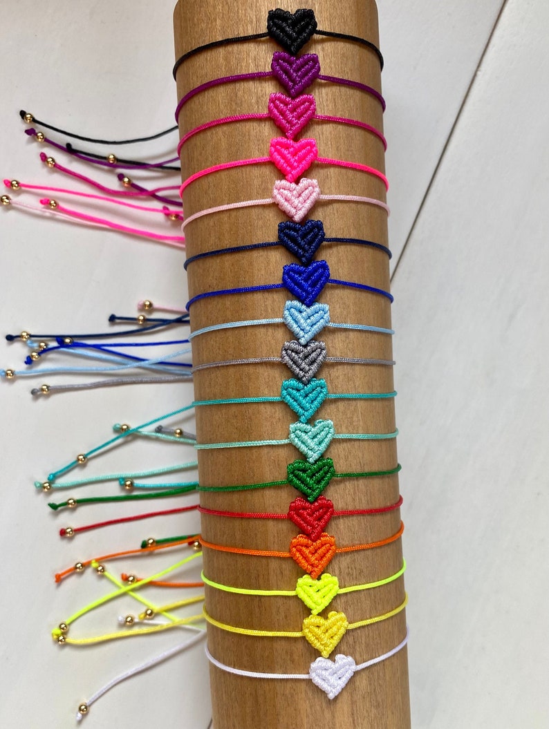 Puede incluir: Una colecci&oacute;n de diecis&eacute;is pulseras de cord&oacute;n ajustables, cada una con un dije de coraz&oacute;n de macram&eacute; en un color diferente, incluyendo negro, morado, rosa, azul, turquesa, verde, rojo, naranja, amarillo y blanco, con peque&ntilde;as cuentas doradas.