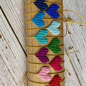 Puede incluir: Una colecci&oacute;n de pulseras hechas a mano con peque&ntilde;os dijes en forma de coraz&oacute;n hechos de hilo tejido apretado en una variedad de colores que incluyen negro, plata, azul marino, azul, celeste, verde, morado, magenta, rosa, rojo, naranja y amarillo.