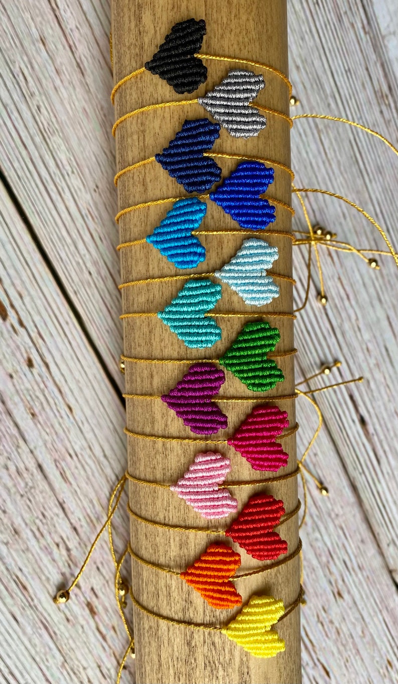 Puede incluir: Una colecci&oacute;n de pulseras hechas a mano con peque&ntilde;os dijes en forma de coraz&oacute;n hechos de hilo tejido apretado en una variedad de colores que incluyen negro, plata, azul marino, azul, celeste, verde, morado, magenta, rosa, rojo, naranja y amarillo.