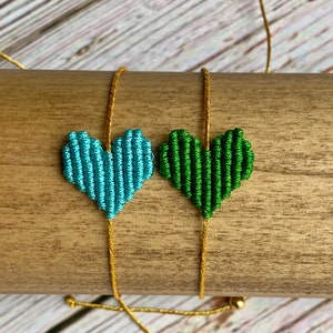 Puede incluir: Dos pulseras de macram&eacute; con dijes en forma de coraz&oacute;n hechos de cord&oacute;n anudado. Un coraz&oacute;n es azul claro y el otro es verde bosque. Ambos dijes est&aacute;n unidos a cordones ajustables de color dorado met&aacute;lico con peque&ntilde;as cuentas doradas en los extremos.