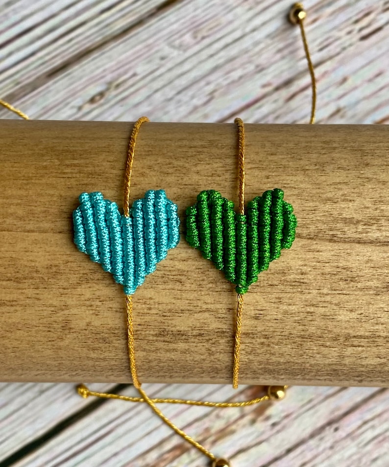 Puede incluir: Dos pulseras de macram&eacute; con dijes en forma de coraz&oacute;n hechos de cord&oacute;n anudado. Un coraz&oacute;n es azul claro y el otro es verde bosque. Ambos dijes est&aacute;n unidos a cordones ajustables de color dorado met&aacute;lico con peque&ntilde;as cuentas doradas en los extremos.