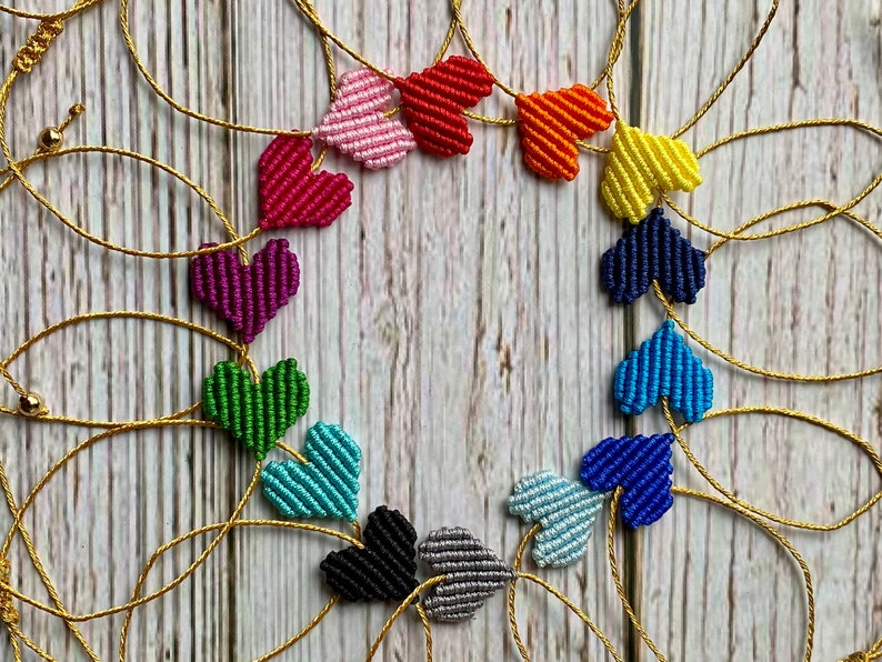 Puede incluir: Una disposici&oacute;n circular de corazones de macram&eacute; en varios colores, incluyendo rosa, rojo, naranja, amarillo, azul oscuro, azul claro, verde, morado, negro y gris. Cada coraz&oacute;n est&aacute; conectado por un cord&oacute;n dorado fino con peque&ntilde;as cuentas doradas.