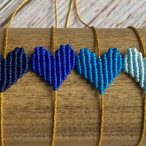 Puede incluir: Cuatro pulseras con dijes en forma de coraz&oacute;n hechos de cord&oacute;n trenzado en un degradado de tonos azules, desde azul marino oscuro hasta azul cielo claro. Cada coraz&oacute;n est&aacute; centrado en un cord&oacute;n dorado met&aacute;lico que sirve como banda ajustable.