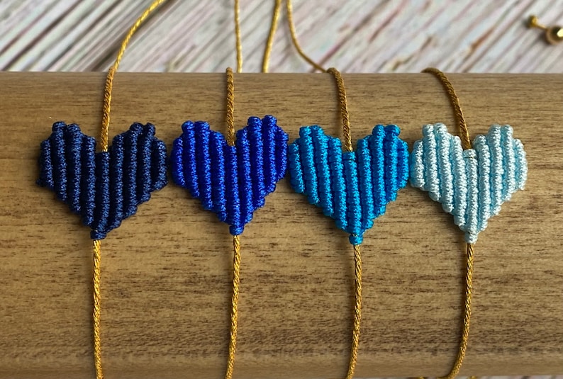 Puede incluir: Cuatro pulseras con dijes en forma de coraz&oacute;n hechos de cord&oacute;n trenzado en un degradado de tonos azules, desde azul marino oscuro hasta azul cielo claro. Cada coraz&oacute;n est&aacute; centrado en un cord&oacute;n dorado met&aacute;lico que sirve como banda ajustable.