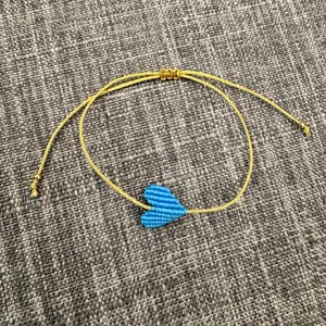 Puede incluir: Una pulsera que presenta un dije de coraz&oacute;n tejido en color azul brillante centrado en un cord&oacute;n fino de color dorado reluciente. El cord&oacute;n incluye un mecanismo de nudo corredizo ajustable y peque&ntilde;as cuentas doradas en los extremos.