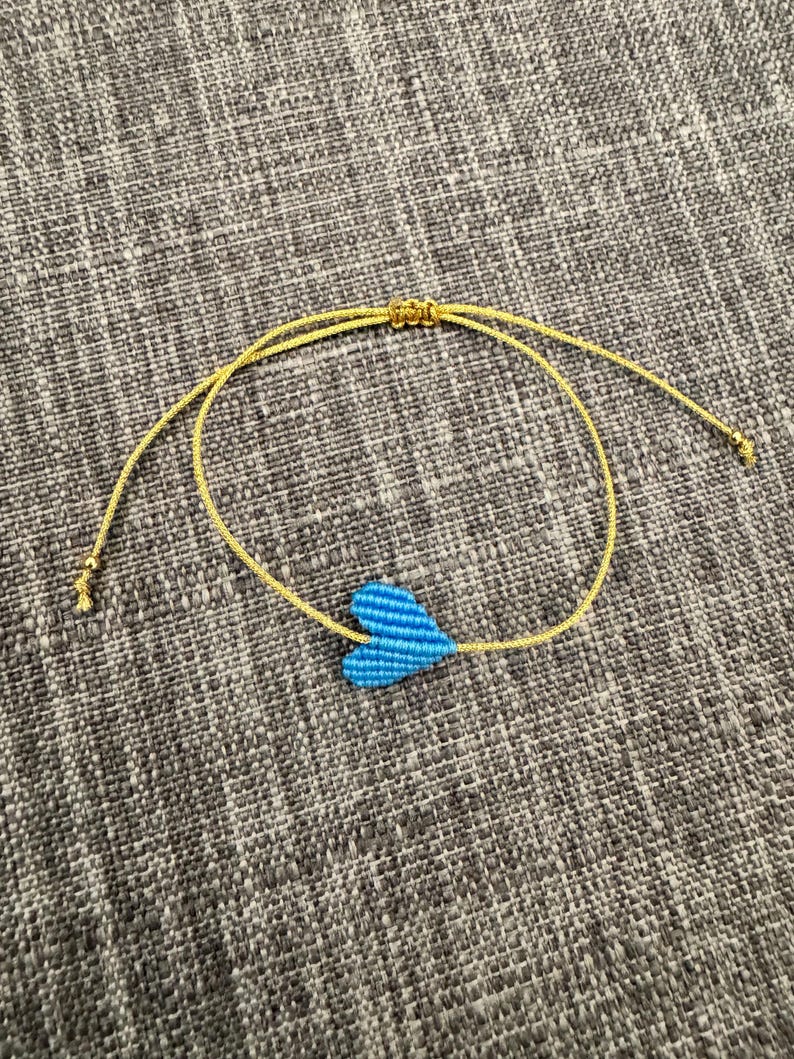 Puede incluir: Una pulsera que presenta un dije de coraz&oacute;n tejido en color azul brillante centrado en un cord&oacute;n fino de color dorado reluciente. El cord&oacute;n incluye un mecanismo de nudo corredizo ajustable y peque&ntilde;as cuentas doradas en los extremos.